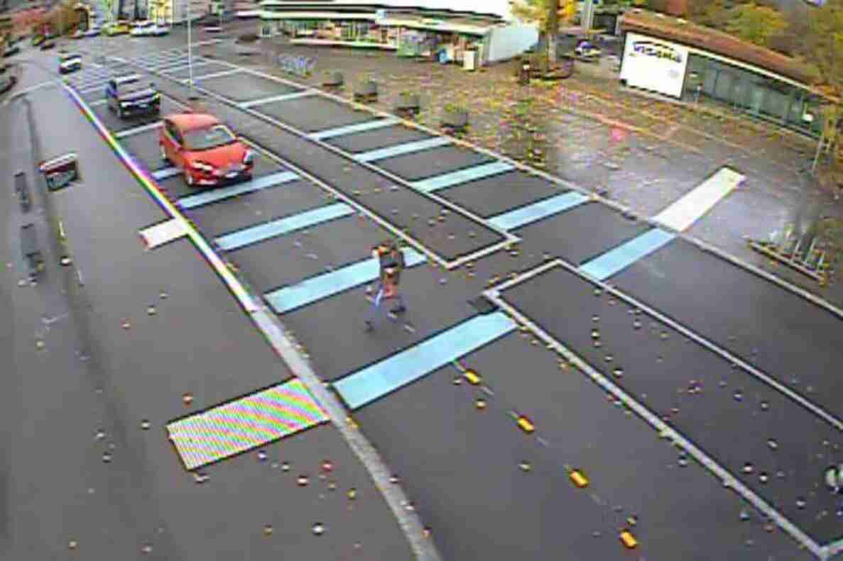 Übersichtsaufnahme einer Kreuzung im Dorfzentrum Münsingen mit testweise blau markierten Querungszonen für Fussgänger auf der Fahrbahn, ergänzt durch orange Leitkegel und Markierungspunkte. Eine Person überquert die Strasse in einer der markierten Zonen, während ein rotes Auto und ein Lieferwagen auf der Fahrbahn warten. Im Hintergrund liegen Parkplätze und Geschäfte.