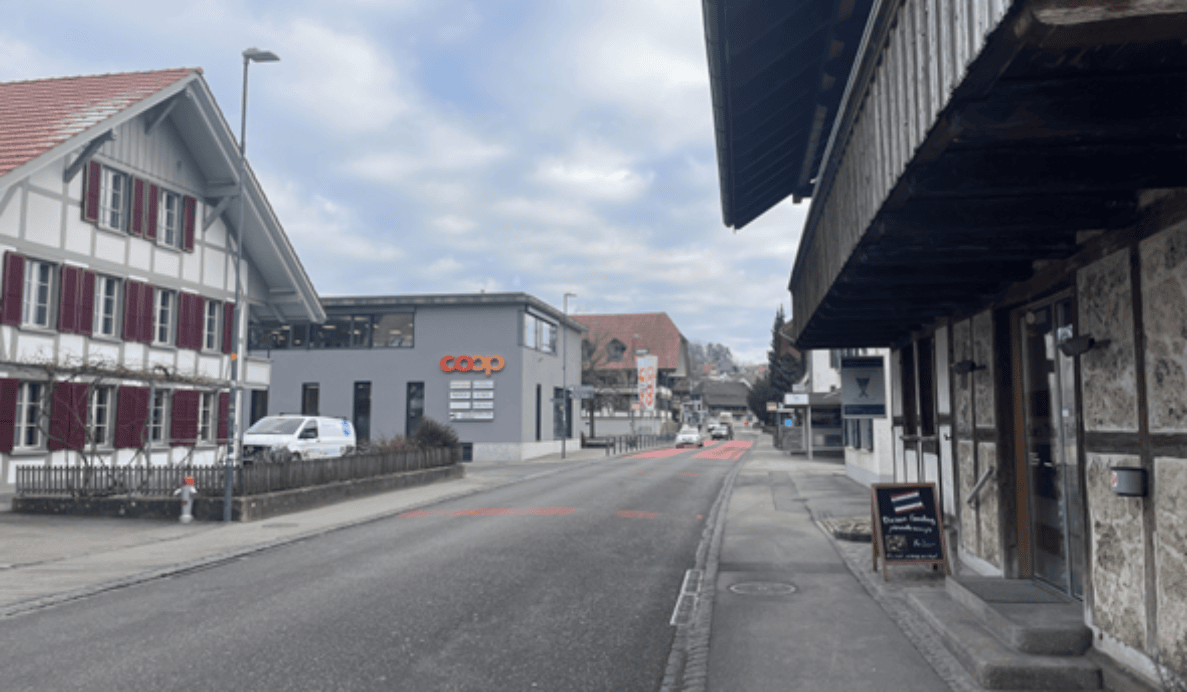 Dorfstrasse in Neuenegg, Blickrichtung Nordost, gesäumt von traditionellen Fachwerkhäusern und modernen Geschäftsbauten mit Coop-Filiale. Zweispurige Fahrbahn mit roten Radstreifen-Markierungen, schmale Trottoirs beidseitig. Ein weisser Lieferwagen parkiert am Strassenrand, ein Fahrzeug fährt talwärts. Weiter hinten sind Fussgängerstreifen sowie weitere Verkehrsteilnehmer erkennbar. Strassenlaternen und Signalisationen deuten auf eine Ortsdurchfahrt mit gemischter Nutzung hin.