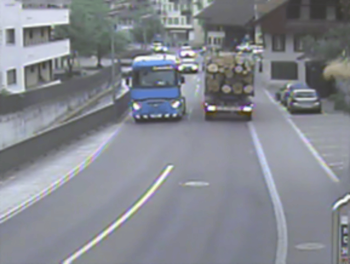Hauptstrasse K248 in Niederlenz mit Gegenverkehr: Ein blauer Lastwagen fährt talwärts, während ihm ein mit Baumstämmen beladener Lastwagen sowie mehrere Personenwagen entgegenkommen. Die zweispurige Strasse ist mit Sperrfläche und Leitlinien markiert, beidseitig verlaufen Trottoirs. Rechts stehen Wohn- und Geschäftshäuser sowie parkierte Fahrzeuge, im Hintergrund ist bewaldetes Gelände sichtbar.