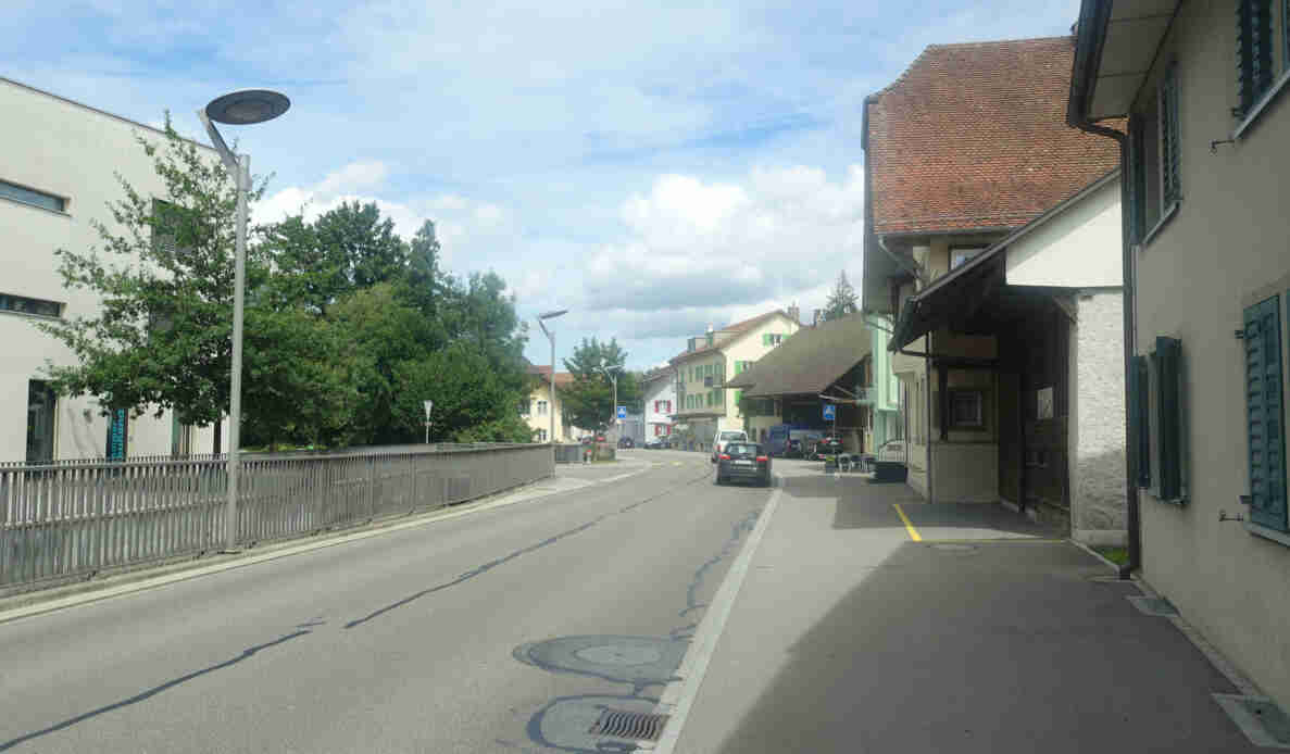 Hauptstrasse in Niederlenz mit Blick über eine Brücke Richtung Dorfzentrum. Am Strassenrand parkierte Fahrzeuge, rechts ein Fussgängerstreifen mit entsprechendem Signal. Die Fahrbahn ist asphaltiert und in gutem Zustand, seitlich verläuft ein Trottoir. Die Bebauung besteht aus historischen Bauernhäusern mit Satteldächern sowie neueren Gebäuden. Keine Personen im Strassenverkehr sichtbar.