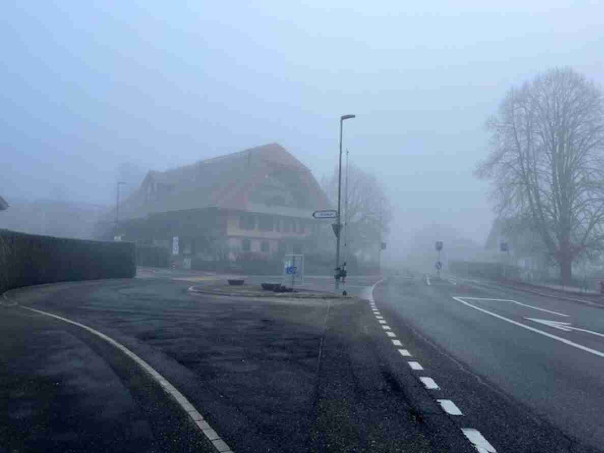 Knoten Oberlindach mit Einmündung von Lindachstrasse und Diemerswilstrasse. Eine kleine Verkehrsinsel trennt die Fahrspuren, auf der Fahrbahn sind Richtungspfeile aufgemalt. Ein Wegweiser für Fahrradverkehr steht am Strassenrand, daneben ein grosses Fachwerkhaus mit rotem Ziegeldach. Fahrbahn und Bankette sind trocken, kein Verkehr sichtbar.