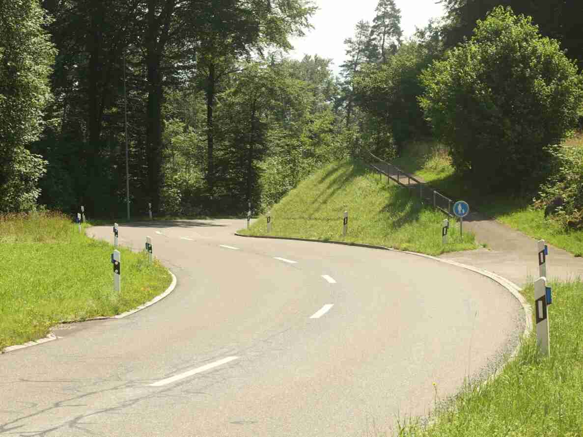 Kurvenreiche, mit Leitpfosten gesäumte Nebenstrasse am Ortsausgang Chrüzlen, gut asphaltiert und ohne Fahrzeuge oder Personen. Rechts zweigt ein separater Fussweg ab, gekennzeichnet mit dem blauen Signal für Fussgänger mit Kind. Im Hintergrund führt ein Steg mit Geländer über die Böschung durch den Wald. Keine Verkehrsmessgeräte sichtbar.