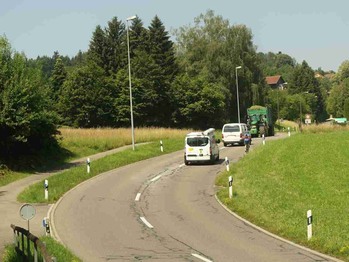 Chrüzlenstrasse in Blickrichtung Westen: leicht kurvige Überlandstrasse mit Mittellinie und rissigem Belag. Zwei weisse Lieferwagen, ein Traktor mit Anhänger und eine radfahrende Person hintereinander in Fahrtrichtung. Am rechten Fahrbahnrand Tempo-60-Signal, Ortstafel und ein Vortrittssignal. Links ein Radweg entlang der Strasse, umgeben von Wiesen und Bäumen.