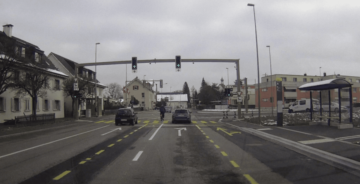 Kreuzung Rheinstrasse/Hauptstrasse mit signalgeregelten Fahrspuren, Ampeln zeigen Grün für Geradeaus und Rechtsabbiegen. Zwei Personenwagen und eine Velofahrperson warten vor der Kreuzung, Fahrbahn ist trocken mit Schneeresten am Fahrbahnrand. Rechts eine Bushaltestelle mit überdachtem Wartebereich, im Hintergrund Wohn- und Gewerbegebäude sowie ein Kirchturm.