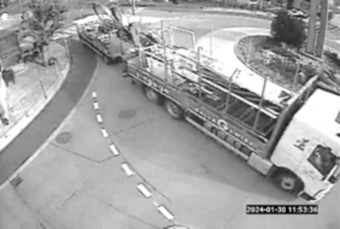Überwachungskamera-Aufnahme in Schwarz-Weiss beim Kreisverkehr Ramsei Bahnhof: Ein Lastwagen mit Anhänger, beladen mit Fahrzeugen respektive Maschinen, biegt auf der Strasse in Richtung Kreisverkehr ab. Die Fahrbahn ist mit einer unterbrochenen Leitlinie markiert, am Strassenrand links verläuft ein Gehweg mit Absperrgitter. Zeitstempel unten rechts: 2024-01-30 11:53:36.