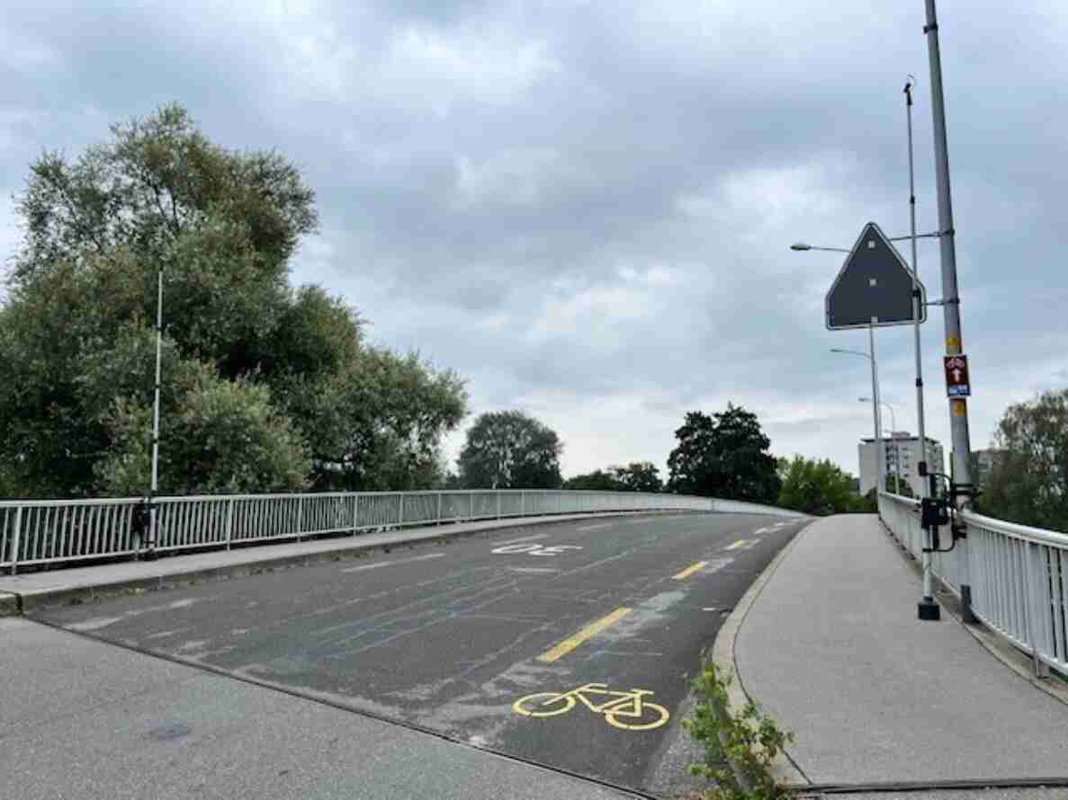 Messquerschnitt in Nidau: Brücke mit metallenem Geländer, auf der Fahrbahn ein gelbes Velo-Piktogramm markiert einen Radstreifen. An einem Mast ist ein Gerät zur Velozählung montiert, daneben ein Vorwegweiser-Schild. Trottoir und Fahrbahn sind baulich getrennt, keine Personen oder Fahrzeuge sichtbar.
