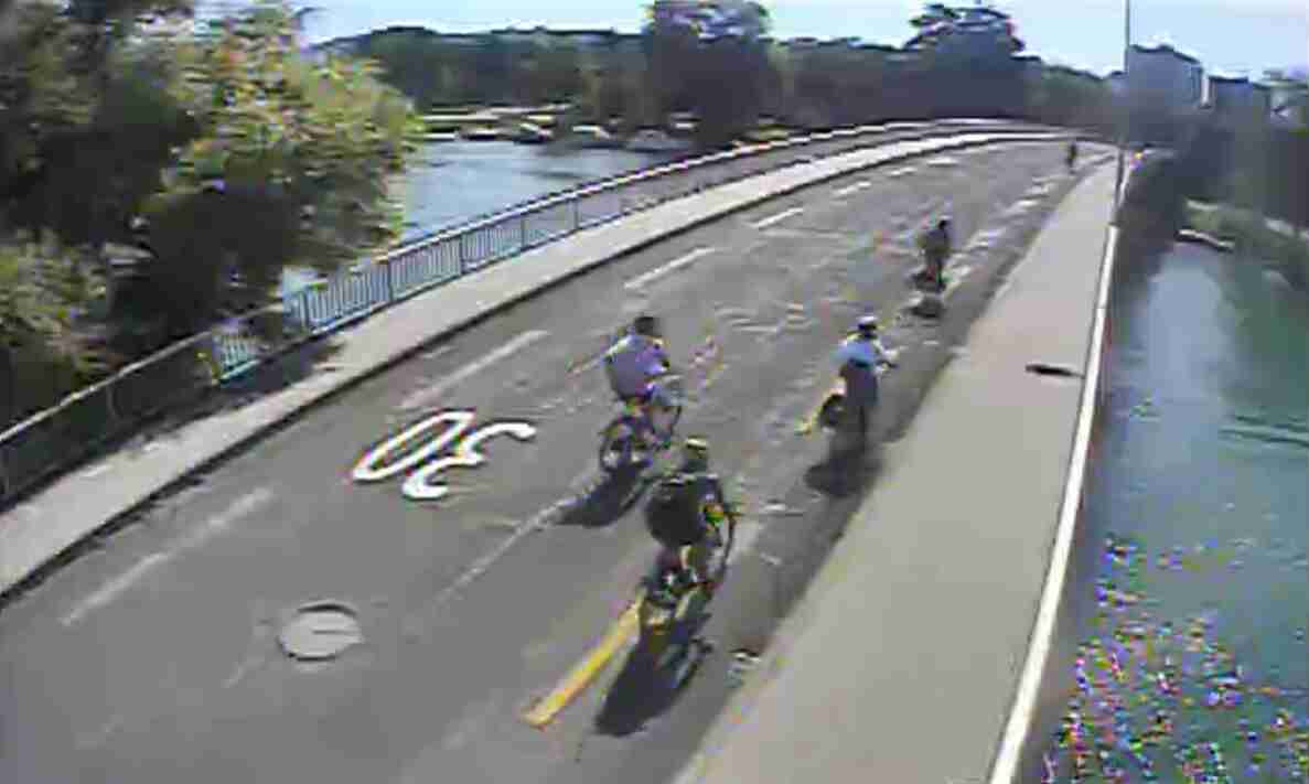 Kamerastandbild einer Brücke über einen Fluss mit separat markiertem Rad- und Fussweg, erkennbar an aufgemalten Velosymbolen auf dem Belag. Mehrere Velofahrende sind hintereinander unterwegs, dazwischen bewegen sich Fussgängerinnen und Fussgänger auf dem Weg. Der Strassenbelag ist trocken und in gutem Zustand. Ein Kanaldeckel ist in der Fahrbahn sichtbar.