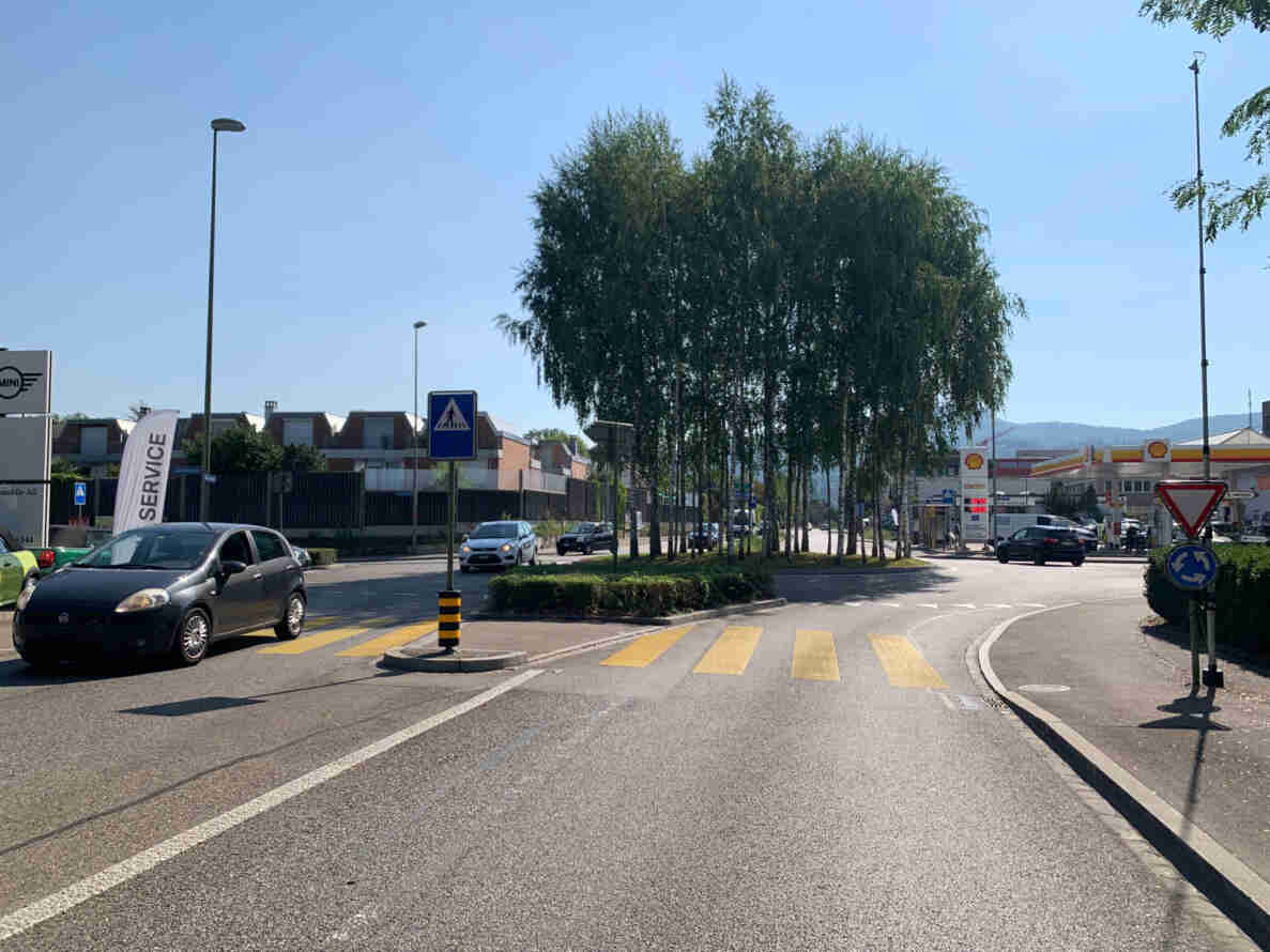 Kreisverkehr in Reinach mit Blickrichtung Westen, umgeben von schlanken Birken. Gelbe Fussgängerstreifen führen zur Kreiselinsel, ein Signal «Kreisverkehr» sowie ein Kein-Vortritt-Dreieck stehen am Fahrbahnrand. Fahrzeuge befahren die Kreiselzufahrten, im Hintergrund eine Shell-Tankstelle und ein Mini-Autohaus. Der Strassenbelag ist trocken und in gutem Zustand, Verkehrsaufkommen moderat.