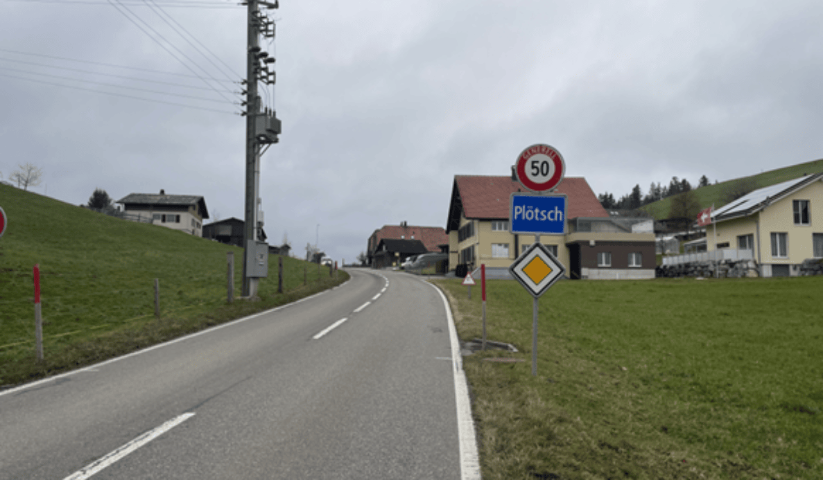 Schulweg Rüti bei Riggisberg mit Sicht Richtung Plötsch: Schmale Überlandstrasse mit Mittelmarkierung und leichtem Gefälle, beidseitig Grünstreifen ohne separaten Fussweg. Rechts Ortstafel Plötsch mit Höchstgeschwindigkeit 50 km/h sowie ein Signal für Strassen mit Vortritt. Vereinzelte Wohnhäuser säumen die Strasse, keine Verkehrsteilnehmenden sichtbar.