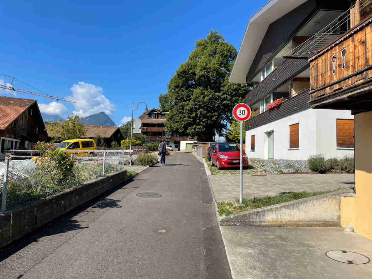 Strasse innerorts mit Signal «Höchstgeschwindigkeit 30» am rechten Fahrbahnrand vor einem Wohnhaus mit Holzfassade und Balkonen. Der Belag ist intakt und asphaltiert, am Strassenrand ein rotes Auto sowie ein gelber Lieferwagen parkiert. Eine Person geht auf dem schmalen Gehweg entlang der Häuserzeile. Im Hintergrund traditionelle Chalets, ein Baukran und ein Berg vor blauem Himmel.