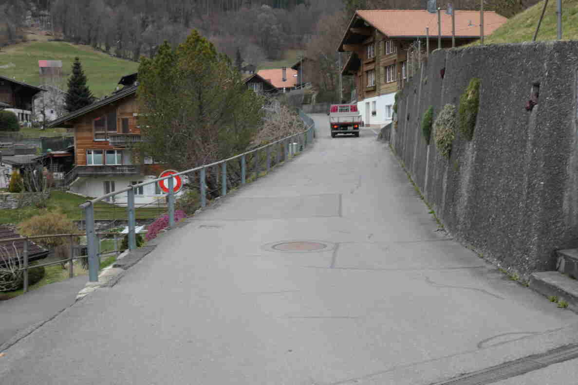 Ansteigende Quartierstrasse in Ringgenberg mit rissigem Asphalt, gesäumt von einem Metallgeländer links und einer Stützmauer rechts. Ein Verbotssignal für Einfahrt verboten steht am Wegrand. Im Hintergrund fährt ein rot-weisser Kleintransporter bergaufwärts an Chalets mit Holzfassaden vorbei, umgeben von Wiesen und Wald am Hang.