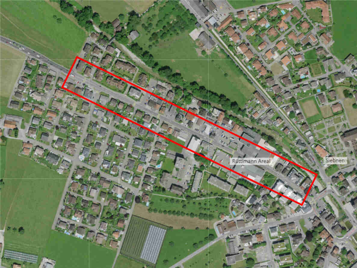 Luftbild von Siebnen mit rot markiertem Perimeter der Ortsdurchfahrt West entlang der durchs Dorf führenden Hauptstrasse. Eingezeichnet ist das Ruttimann Areal sowie umliegende Wohnhäuser, Gärten und Nebenstrassen. Die Strasse verläuft diagonal durch den markierten Bereich und verbindet die Ortseinfahrt im Westen mit dem dichter bebauten Ortskern gegen Osten.