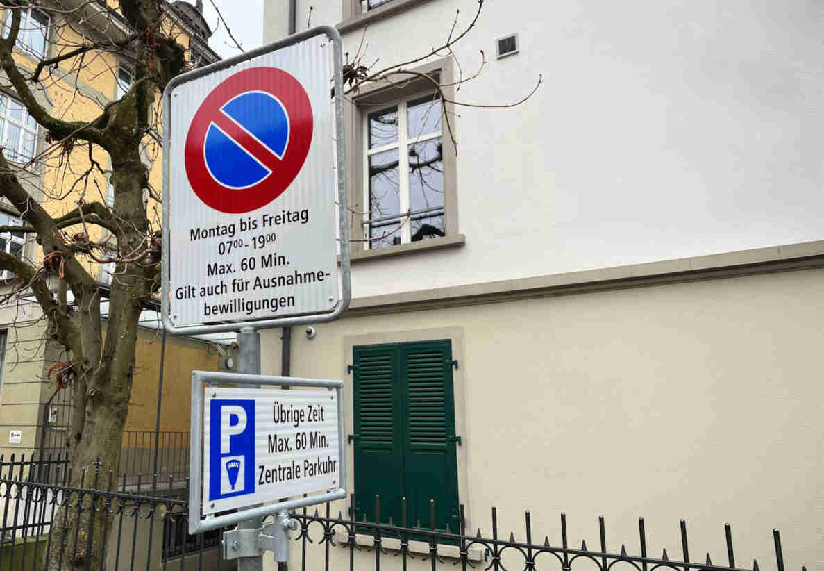 Verkehrsschild «Parkieren verboten» mit Zusatztafeln «Montag bis Freitag 07.00–19.00, Max. 60 Min., gilt auch für Ausnahmebewilligungen» und darunter «Übrige Zeit Max. 60 Min., Zentrale Parkuhr». Die Signalisation regelt die temporäre Nutzung der Parkfelder. Im Hintergrund ein Wohnhaus mit grünen Fensterläden, ein kahler Baum sowie ein schmiedeeisernes Gartengitter.