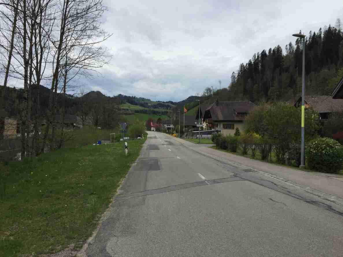 Gerade Dorfstrasse mit Mittellinie und leicht rissigem Belag, gesäumt von einem gepflasterten Trottoir. Links steht ein Vortrittssignal am Strassenrand, im Hintergrund ein Wegweiser sowie eine Schweizer Fahne bei einem traditionellen Bauernhaus mit Ziegeldach. Umgeben von Wiesen, Bäumen und bewaldeten Hügeln, keine Fahrzeuge oder Personen auf der Strasse sichtbar.