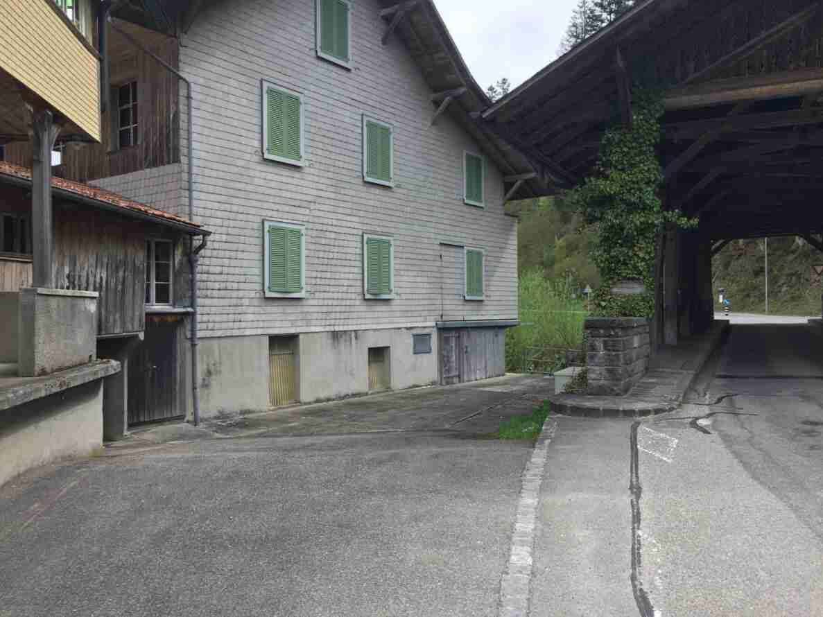 Schindelverkleidetes Bauernhaus mit grünen Fensterläden, daneben führt eine überdachte Holzbrücke über einen Bach. Die schmale Zufahrtsstrasse mit Kopfsteinpflaster-Randstein verengt sich zur Brücke, am Ende sind ein Vortrittssignal und ein Fahrverbotsschild für Motorfahrzeuge erkennbar. Der Belag ist trocken und teils rissig, keine Personen sichtbar.