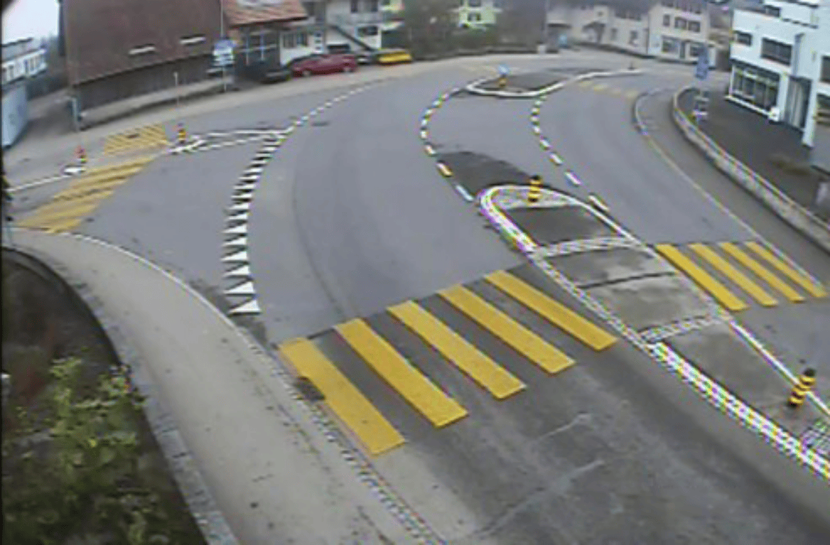 Standbild einer Videokamera zeigt den Knoten Hauptstrasse/Poststrasse/Unterdorfstrasse in Utzenstorf mit einer Verkehrsinsel und provisorischer Verkehrsführung. Gelbe Fussgängerstreifen queren die breite Fahrbahn, weisse und schwarze Leitmarkierungen sowie orange Leitkegel begrenzen den Verkehrsraum. Parkierte Fahrzeuge stehen am Strassenrand, Gebäude säumen die Kreuzung. Keine Personen oder Fahrzeuge sind auf der Fahrbahn unterwegs.