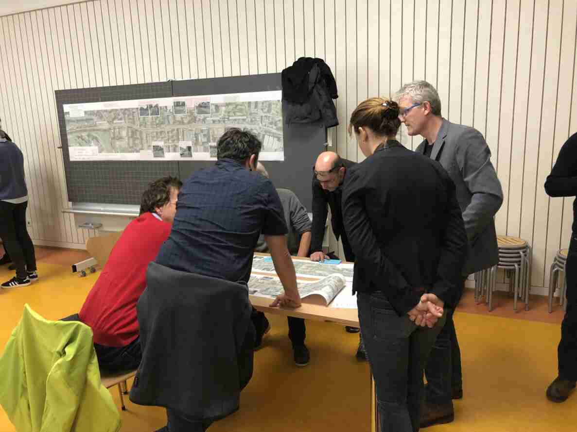 Gruppe von Personen in einem Sitzungszimmer, die um einen Tisch versammelt sind und einen ausgerollten Plan mit Strassenkarte und Verkehrssituationsfotos betrachten. Im Hintergrund hängt eine grosse Übersichtstafel mit Luftaufnahme und Strassenfotos an einer Pinnwand. Eine gelbe Warnjacke liegt über einem Stuhl im Vordergrund.