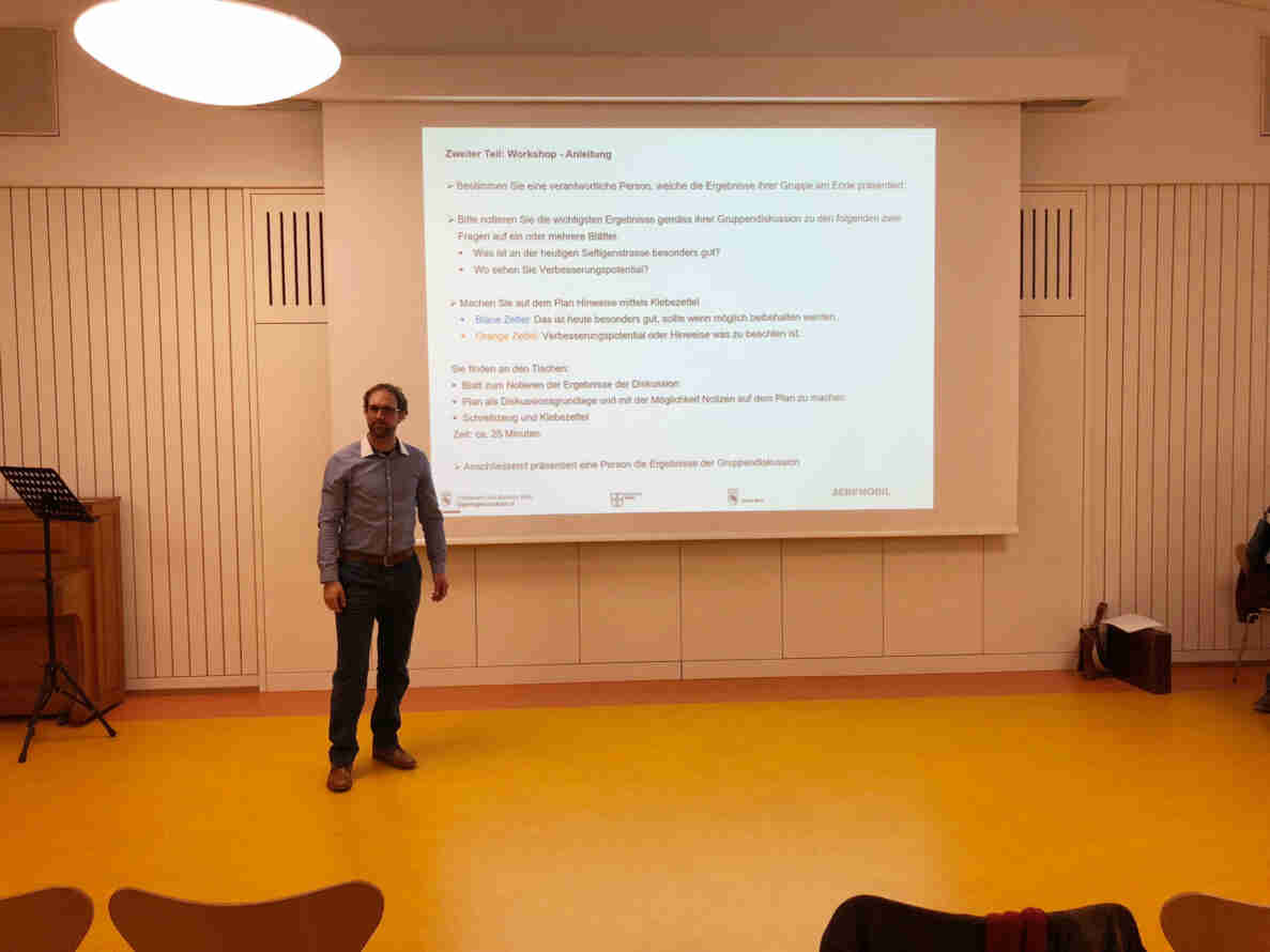 Person steht vor einer Projektionsleinwand in einem Saal mit gelbem Boden und erklärt eine Workshop-Anleitung zur Seftigenstrasse. Die Präsentation enthält Fragen zu positiven Aspekten und Verbesserungspotential der Strasse sowie Anweisungen zur Gruppendiskussion mit farbigen Klebezetteln. Logos von Tiefbauamt des Kantons Bern, Gemeinde Köniz, Stadt Bern und Bernmobil sind unten sichtbar. Im Vordergrund stehen leere Stühle.