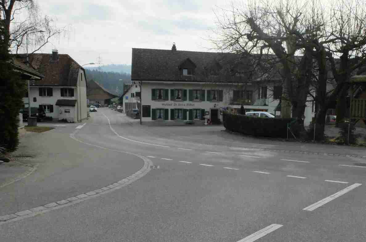 Blick auf die Oltenstrasse Richtung Westen mit Kurvenführung durch aufgemalte Leitlinien. Im Hintergrund der Gasthof St. Urs u. Viktor, davor parkierte Fahrzeuge sowie ein Lieferwagen der Verkehrsteiner AG am Strassenrand. Die trockene Fahrbahn ist mit Sicherheitslinien markiert, Vorfahrtsregelung ist durch ein Signal an der Kreuzung erkennbar. Keine Personen im Strassenraum sichtbar.