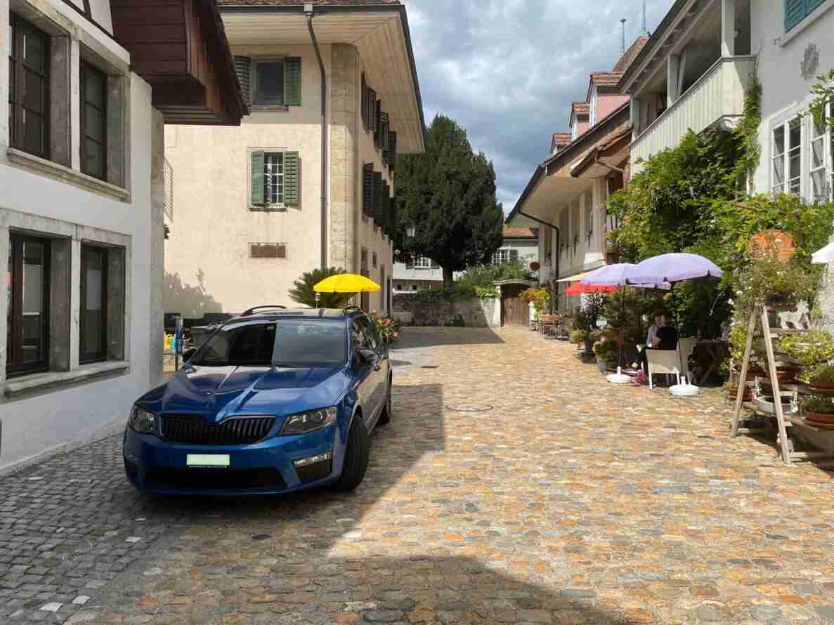 Kopfsteinpflastergasse in einer Schweizer Altstadt ohne Verkehrssignale, was auf eine verkehrsberuhigte Zone oder Fussgängerbereich hindeutet. Ein blauer Kombi steht am Strassenrand vor historischen Gebäuden mit grünen Fensterläden. Rechts sitzen zwei Personen unter bunten Sonnenschirmen vor einem Café, umgeben von Blumentöpfen und Pflanzen an den Fassaden. Der Boden besteht aus unebenem, gepflegtem Pflasterstein.