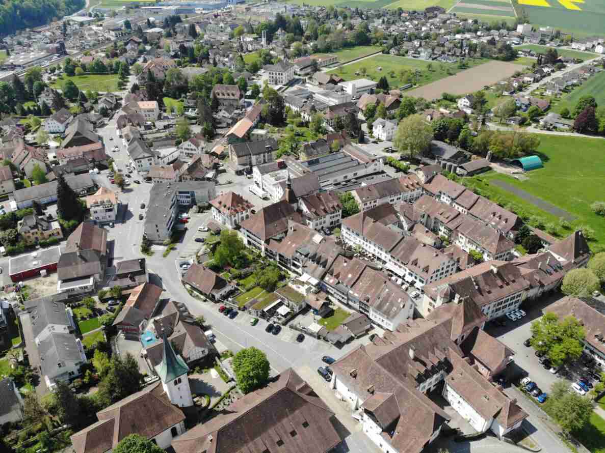 Luftaufnahme des «Städtli» Wangen an der Aare mit dichter Bebauung aus Häusern mit Ziegeldächern, einem Kirchturm mit grüner Spitze sowie umliegenden Feldern. Mehrere Strassen durchziehen den Ortskern, an den Fahrbahnrändern parkieren zahlreiche Fahrzeuge. Die Strassen sind in gutem Zustand und weisen Mittellinien sowie Parkfeldmarkierungen auf, was auf innerörtliche Erschliessungsstrassen hinweist. Personen sind auf den Strassen keine erkennbar.