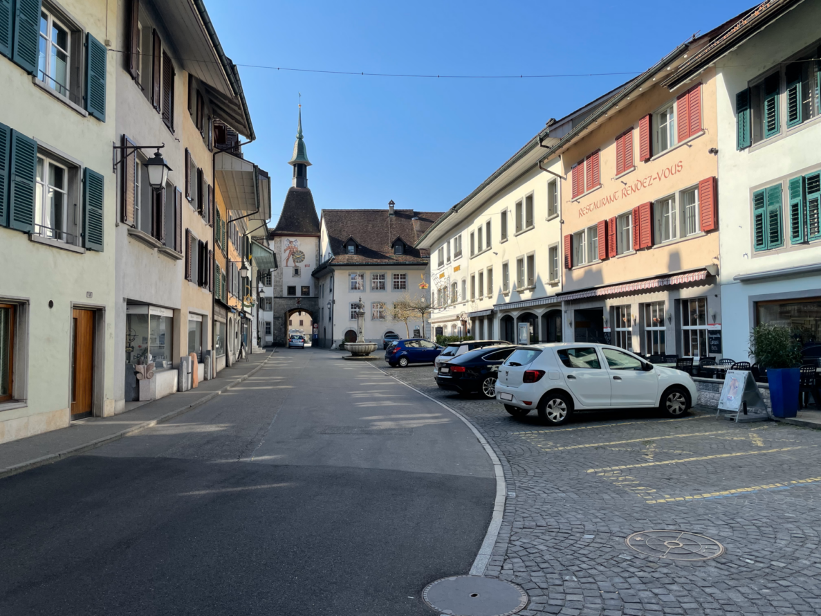Gemeindestrasse in der Altstadt von Wangen an der Aare mit Blick auf den Turm mit Uhr und Fresko. Rechts sind Fahrzeuge auf gepflasterten Parkfeldern vor dem Restaurant Rendez-vous abgestellt, links Wohn- und Geschäftshäuser mit farbigen Fensterläden. Die Strasse verläuft asphaltiert und leicht kurvig, ohne sichtbaren Verkehr oder Personen, ein Brunnen steht vor dem Torbogen.