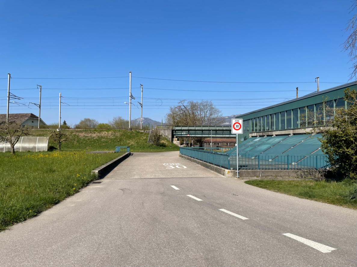 Gemeindestrasse in Wangen an der Aare mit Signal «Zone 30» und entsprechender Bodenmarkierung. Die schmale Fahrbahn ohne Trottoir führt über eine Brücke mit türkisfarbenem Geländer, rechts ein Gebäude mit schräg verglastem Vordach. Im Hintergrund eine Fussgängerbrücke über Bahngleise mit Fahrleitungsmasten, darauf zwei Velofahrende. Wiesen säumen die Strasse beidseitig.