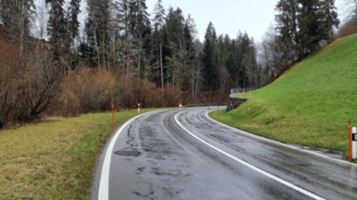 Kurvenbereich KS 11 in Weissenburg, Blickrichtung West: Nasse, kurvige Strasse mit Mittellinie und Randmarkierungen, gesäumt von Leitpflöcken mit orangen Reflektoren. Keine Fahrzeuge oder Personen sichtbar, kein Gegenverkehr erkennbar. Fahrbahn zweispurig, Bankette teils grasbewachsen. Rechts eine Leitplanke im Kurvenverlauf, keine sichtbaren Verkehrssignale oder Messgeräte.