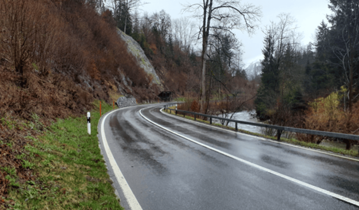 Kantonsstrasse 11 westlich von Weissenburg, Blickrichtung Weissenburg. Nasse, kurvige zweispurige Strasse entlang eines Flusses, links durch Fels begrenzt, rechts Leitplanke zum Gewässer hin. Keine Fahrzeuge oder Personen sichtbar, Fahrbahnmarkierung deutlich erkennbar, Leitpfosten am Strassenrand.