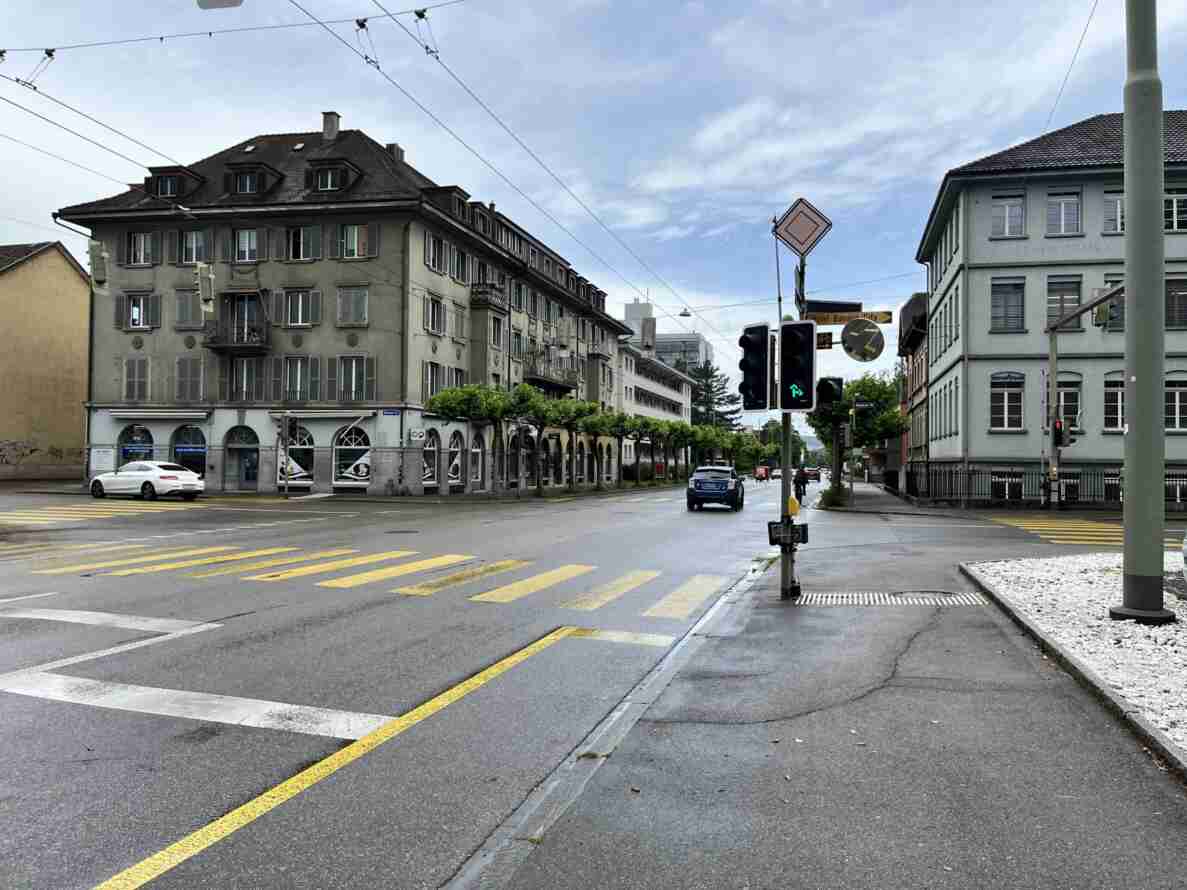 Strassenkreuzung Neuwiesenstrasse/Wülflingerstrasse mit nasser Fahrbahn und gelb-weisser Fussgängerstreifen-Markierung. Ampel zeigt Grün für Rechts- und Linksabbiegepfeile, daneben Verkehrsspiegel und Wegweiser «Hotel Banana City». An der Ampelmaste sind Messgeräte zur Verkehrsmessung montiert. Ein Auto fährt geradeaus, eine Velofahrperson nähert sich der Kreuzung. Historische mehrstöckige Wohn- und Geschäftshäuser säumen die Strasse, Fahrleitungen überspannen die Fahrbahn.