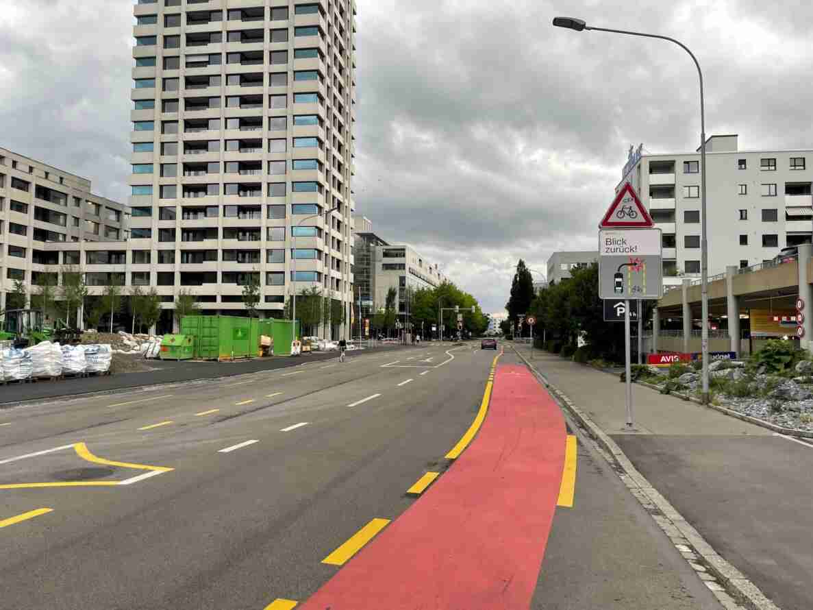 Mehrspurige Stadtstrasse mit rot markiertem Radweg entlang gelb unterbrochener Linien. Rechts Warntafel «Achtung Velo» mit Zusatztafel «Blick zurück!» zur Rechtsabbiegesituation, darunter Parkschild mit 4h-Beschränkung. Im Hintergrund Ampeln mit Tempo-30-Zone, eine radfahrende Person, ein Auto sowie Hochhäuser und eine Baustelle mit Baucontainern am Strassenrand.