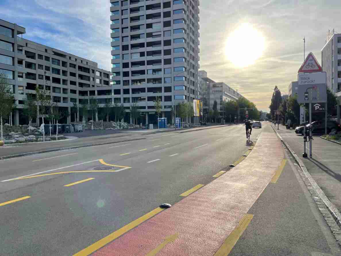 Breite Strasse in Neubauquartier mit rot markiertem Radweg, auf dem eine Person Velo fährt. Rechts ein Warnschild «Blick zurück!» mit Piktogramm für Fahrrad und Auto sowie ein Gerät zur Messung an einem Mast. Weiter hinten Verkehrsampeln, ein Hinweisschild «P 24h» und weitere Personen zu Fuss. Hochhäuser und mehrstöckige Wohnbauten säumen die Strasse, Fahrbahn trocken und frei von Verkehr.