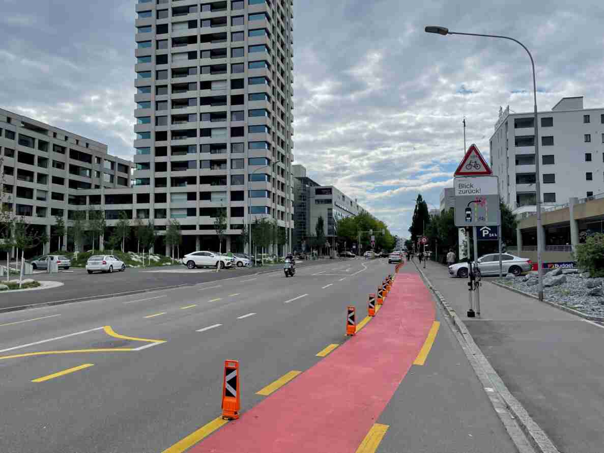 Mehrspurige Stadtstrasse zwischen Hochhäusern mit rot markiertem, durch orange Leitkegel abgetrenntem Radweg. Warnschild «Achtung Velofahrende» mit Zusatztafel «Blick zurück!» weist auf Konfliktbereich zwischen Auto- und Veloverkehr hin, darunter Hinweis auf Parkhaus 24h. Ein Motorroller fährt auf der Fahrbahn, Fussgänger bewegen sich auf dem Trottoir, Fahrzeuge parkieren am Strassenrand.