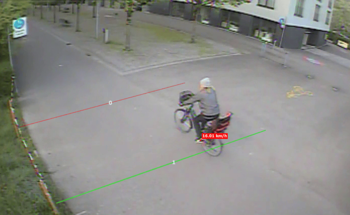 Videostandbild aus der Binzallee in Zürich: Eine radfahrende Person mit heller Mütze und Gepäcktaschen fährt über einen breiten Vorplatz auf ein Wohn-/Geschäftsgebäude zu. Zur Geschwindigkeitsmessung anhand der Videoaufnahmen sind zwei Referenzlinien (rot und grün) mit Nummerierung eingeblendet, eingeblendet wird ein Wert von 16.01 km/h. Am linken Bildrand steht eine blaue Hinweistafel, im Hintergrund sind ein Verkehrsschild sowie parkierte Fahrräder erkennbar.