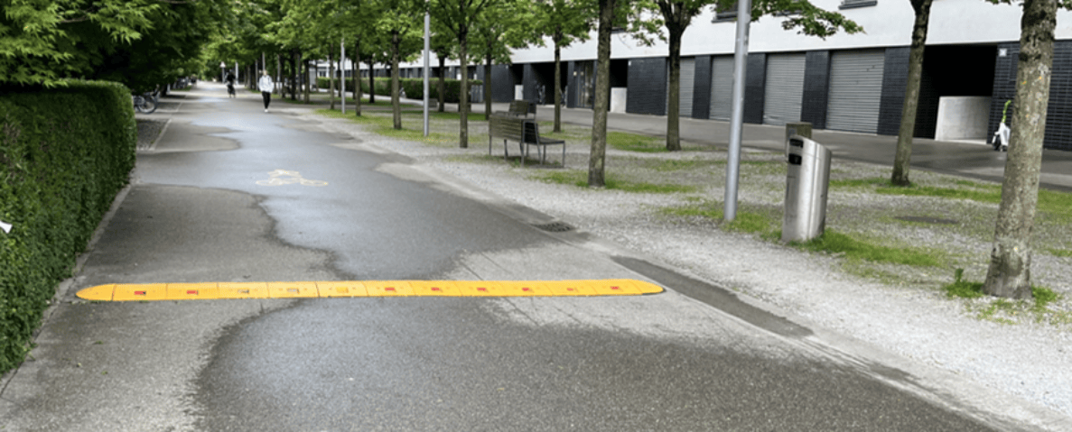 Fuss- und Radweg Binzallee in Zürich mit gelb-roter, temporär installierter Schwelle quer über dem Belag als Geschwindigkeitsbremse. Auf dem Asphalt ist eine Velo-Markierung sichtbar. Am Wegrand stehen Bäume, eine Sitzbank und ein Abfallbehälter, im Hintergrund geht eine Person auf dem Weg, rechts stehen Gebäude mit Garagentoren.