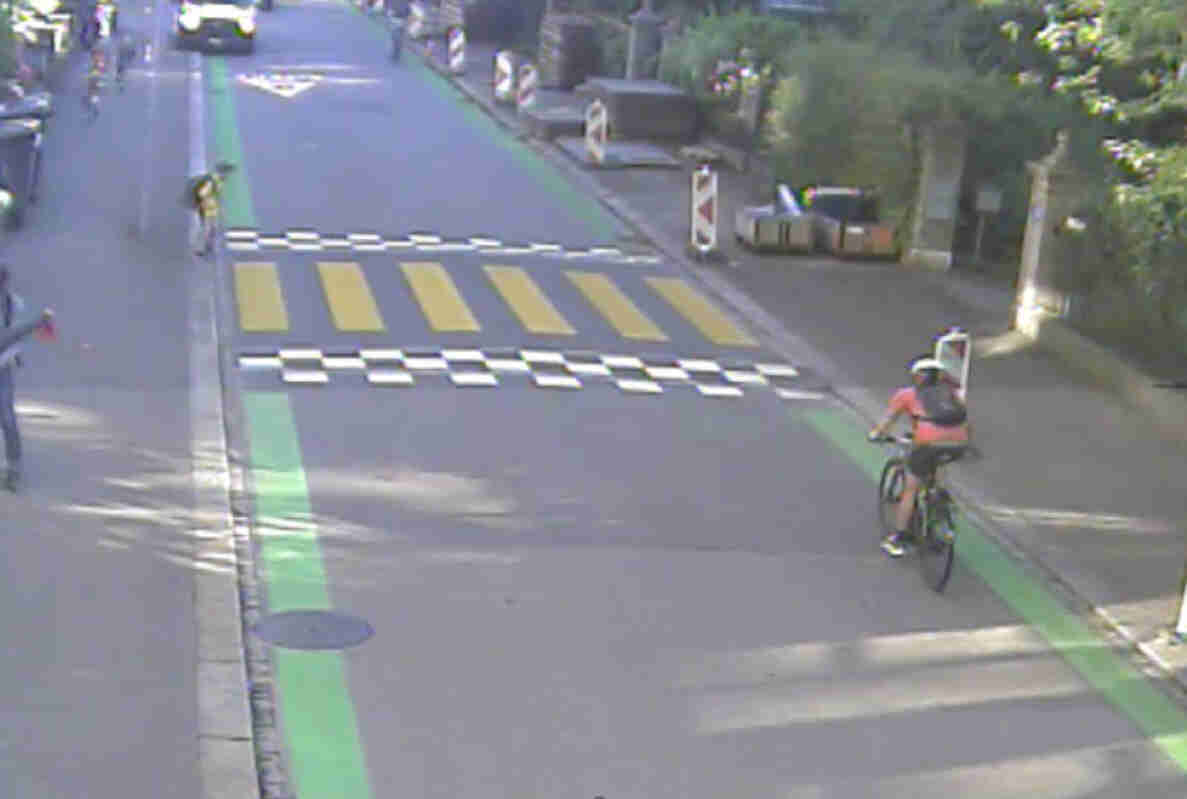 Blick von oben auf die Mühlebachstrasse in Zürich mit grün markierten Radstreifen beidseits der Fahrbahn. Ein Fussgängerstreifen mit gelb-schwarzer Markierung quert die Strasse, davor stehen Baustellensignale. Ein velofahrende Person mit orangem Rucksack nähert sich dem Streifen, während im Hintergrund ein Schulkind mit gelber Mütze am Strassenrand wartet.
