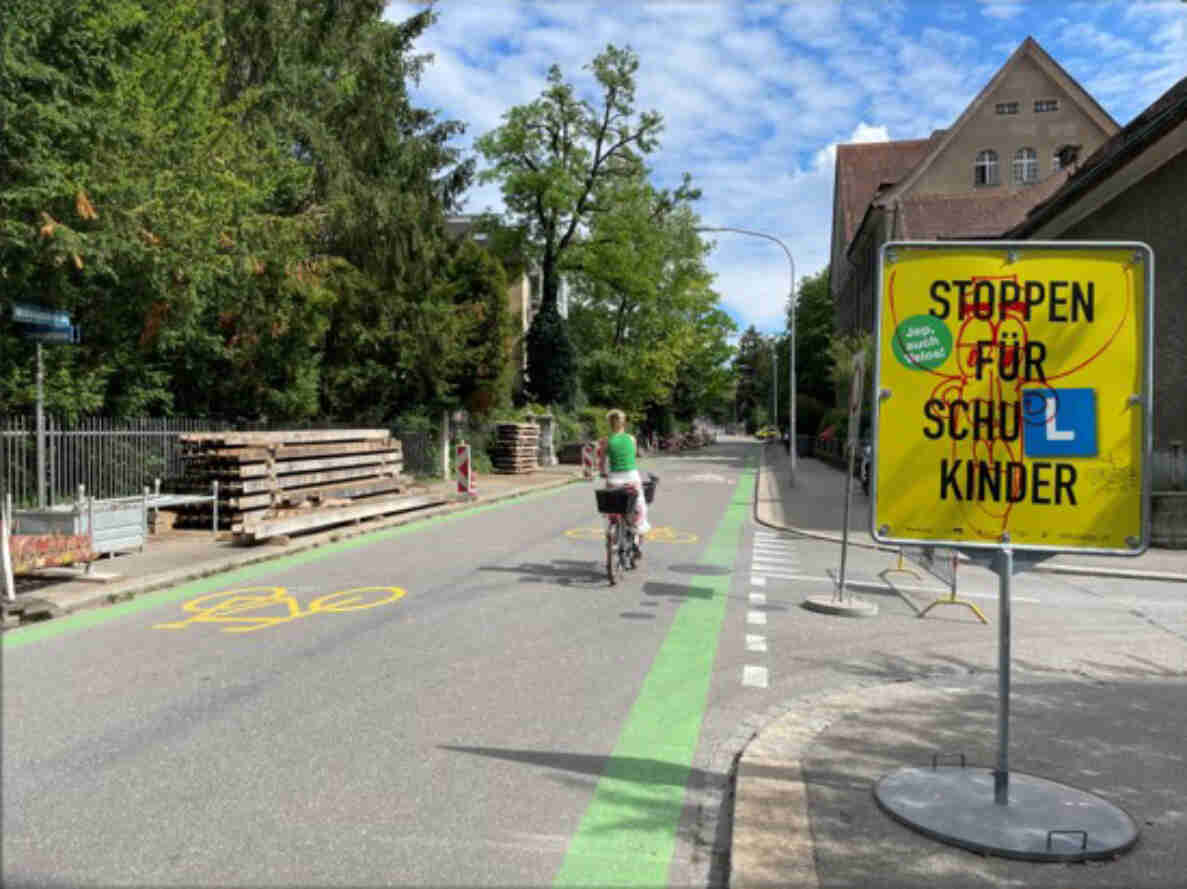 Velovorzugsroute Muehlebachstrasse in Zuerich mit gruen markierten Velostreifen beidseitig der Fahrbahn und aufgemalten Velosymbolen. Eine Person faehrt mit dem Velo auf dem rechten Streifen stadteinwaerts. Rechts steht ein gelbes Warnschild «Stoppen fuer Schulkinder». Links Baumaterial-Lagerplatz hinter einem Bauzaun, dahinter Baeume und ein Gebaeude mit Ziegeldach.
