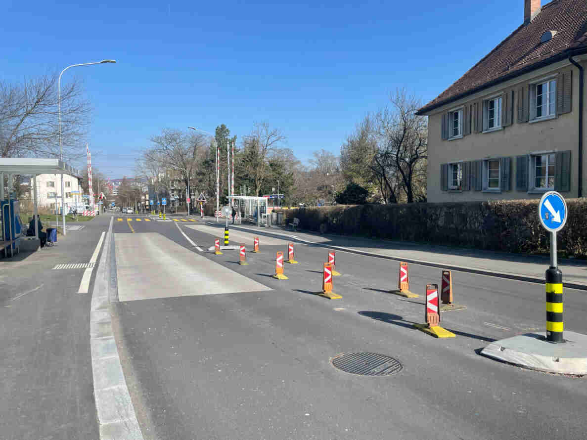 Strasse mit Bushaltestellen beidseits, provisorisch markiert durch rot-weisse Leitkegel und Leitbaken auf einer Mittelinsel mit blauem Rechtsvorschriftssignal. Frischer Asphaltbelag mit Fahrbahnmarkierungen und Fussgängerstreifen. Am Strassenrand warten Personen an der Haltestelle. Im Hintergrund weitere Signalisationen, Bäume ohne Laub und ein mehrstöckiges Wohnhaus mit Fensterläden.