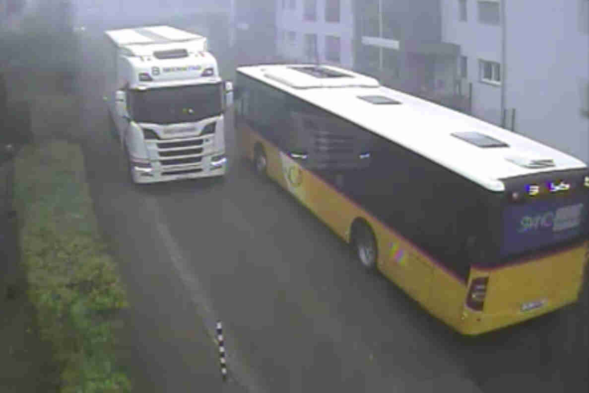 Enge Begegnung eines Lastwagens mit Kühlauflieger und eines gelben Postautos auf der Hinterfeldstrasse in Zwingen. Die Fahrbahn ist schmal, sodass beide grossen Fahrzeuge nur mit geringem seitlichem Abstand aneinander vorbeifahren können. Am Fahrbahnrand ist eine Bodenmarkierung mit Schachbrettmuster sichtbar, links begrenzt eine Hecke die Strasse.