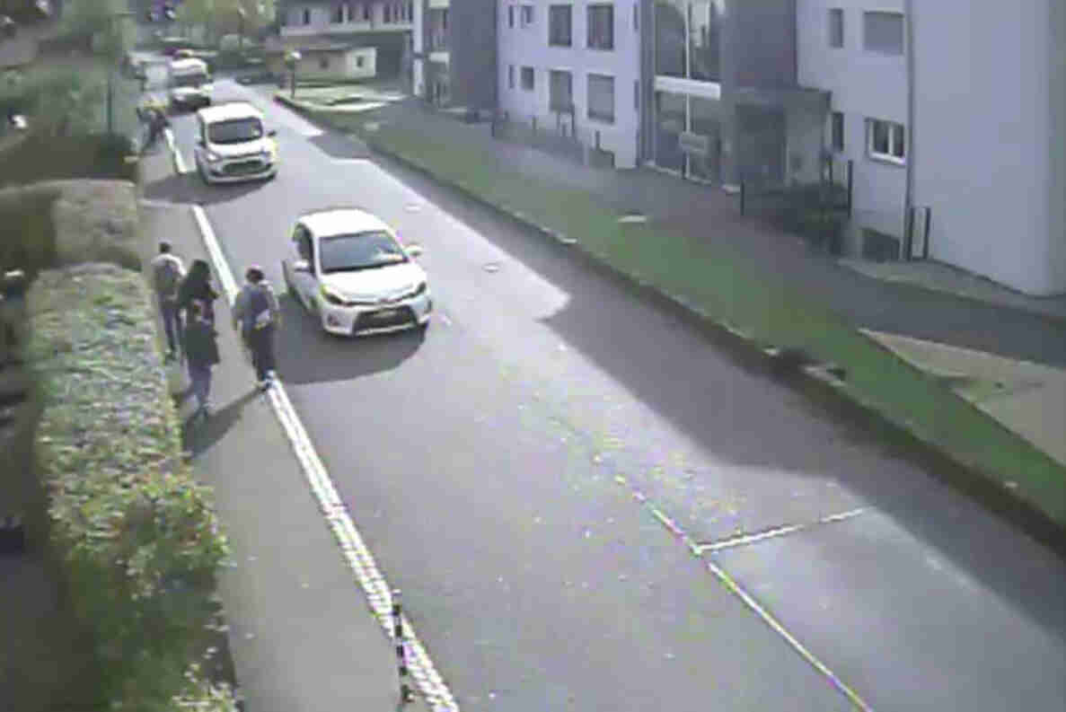 Hinterfeldstrasse in Zwingen: Auf der Fahrbahn fahren mehrere Autos hintereinander, während links auf dem schmalen, einseitigen Trottoir direkt neben der Hecke drei Personen zu Fuss unterwegs sind. Der geringe Abstand zwischen den Fussgängern und den vorbeifahrenden Fahrzeugen zeigt die unzureichende Breite des Trottoirs. Rechts der Strasse befinden sich Mehrfamilienhäuser mit begrünten Vorplätzen.