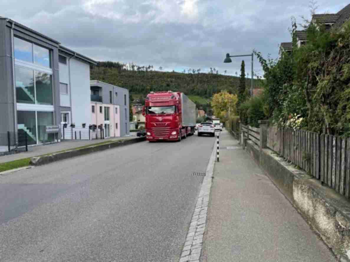 Enge Hinterfeldstrasse in Zwingen mit rotem Lastwagen, der entgegenkommenden Personenwagen entgegenfährt, sodass die Fahrbahn kaum Platz für beide Richtungen bietet. Rechts begleitet ein schmaler Gehweg mit Randstein die Strasse, entlang Holzzäunen und Vorgärten. Links stehen mehrstöckige Wohngebäude direkt an der Strasse. Die Verkehrssituation zeigt die beengten Platzverhältnisse für Fahrzeuge und Fussgänger in diesem Wohnquartier.